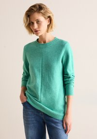 Turkos tröja, rund hals, ribbade mudd och nederkant, mjuk textur, avslappnad passform, kombinerad med blå jeans. Minimalistisk design, inga mönster.