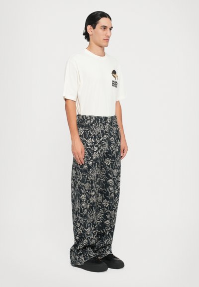 Weißes, oversized T-Shirt mit einem schwarzen Grafikdesign auf der Brust, kombiniert mit lockeren, dunkelblau-beigen Blumenmuster-Hosen, getragen mit schwarzen Schuhen.