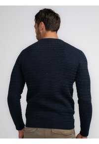 Pulover tricotat bleu marin, cu țesătură texturată, manșete și tiv din bumbac cu nervuri, având un design clasic cu gât rotund. Detalii de cusătură vizibile pe spate.