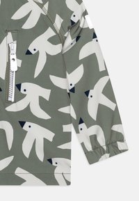 Manica e lato di una giacca verde con motivo di uccelli bianchi, polsino elasticizzato e tasca con zip bianca su sfondo uniforme.