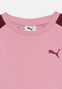 Svetlo roza majica s kratkimi rokavi z maroon aksenti na ramenih. Ima okroglo ovratnico in vezeno maroon Puma logo na prsih. Mehka tekstura tkanine.