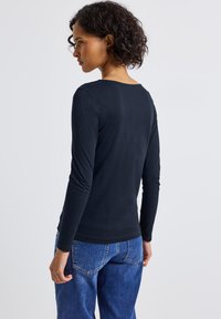 Frau mit kurzen lockigen Haaren, die ein schwarzes Langarmshirt und eine blue Jeans trägt, steht leicht abgewandt vor einem schlichten Hintergrund.