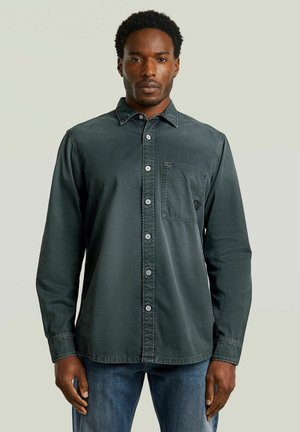 Man met kort krullend haar, gekleed in een donkergroen overhemd met knopen en een borstzak, en een blauwe spijkerbroek, staand tegen een effen lichte achtergrond.