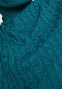 Teal neulepusero, jossa on ribbikuvioitu poolokaulus ja lettokuvio. Pehmeä rakenne, jossa on kohotettua yksityiskohtaa kautta kankaan.