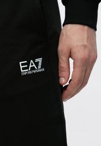 Pantalones joggers negros de corte ajustado, con un logo blanco de EA7 Emporio Armani en el muslo, textura de tela suave y bolsillos laterales.