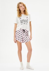 Valkoinen puuvillapaita, jossa on "GOTHIC"-teksti ja kissamaisia piirteitä; vaaleanpunaiset shortsit, joissa on mustia sydänkuvioita; rento kotivaatetus.