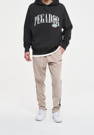 Persoon in zwarte PEGADOR-hoodie, beige trainingsbroek met zwarte strepen en witte sneakers, staand met één hand in de zak.