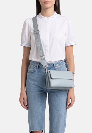 Donna che indossa una camicetta bianca a maniche corte e jeans azzurri, con una borsa a tracolla azzurra con una tracolla larga decorata.