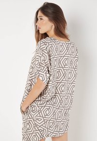 Beige Tunika in lockerer Passform mit geometrischem braunem Muster, kurzen Ärmeln und seitlichen Taschen. Weicher, leichter Stoff mit entspannter Silhouette.