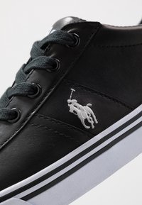 Svart lädersneaker med mörka snören, vit rand på sulan och en vit broderad hästlogotyp på sidan. Texturerad yta.