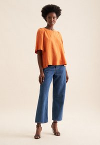 Top in lino arancione con maniche corte, vestibilità ampia; abbinato a jeans in denim blu a vita alta a gamba larga e sandali neri con tacco alto e fasce.
