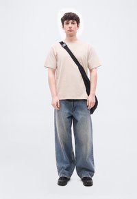 Νεαρός άνδρας που στέκεται φορώντας μπεζ μπλουζάκι, φαρδιά ξεθωριασμένα τζιν, μαύρα αθλητικά παπούτσια και μαύρη τσάντα χιαστί μπροστά σε απλό λευκό φόντο.