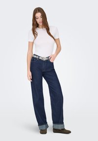 Jean large bleu foncé avec un revers bleu clair, associé à un t-shirt blanc ajusté et une ceinture décorative avec un motif argenté.