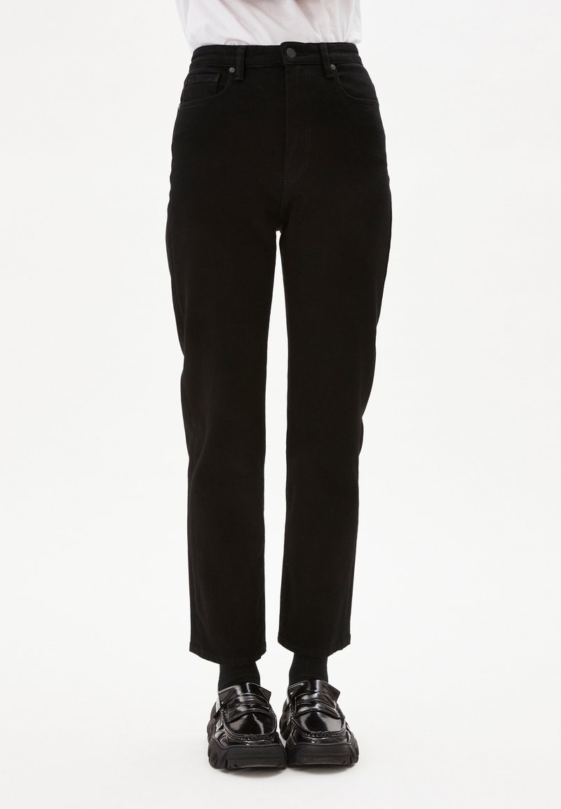 Persona vestida con pantalones negros de talle alto, calcetines negros y mocasines negros brillantes, de pie frente a un fondo blanco.