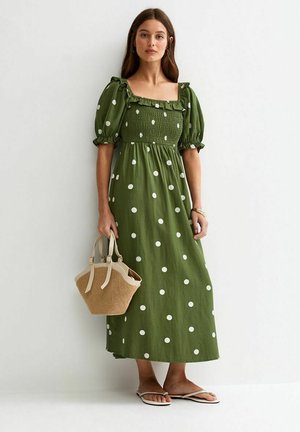 Femme portant une robe midi verte olive à pois blancs et manches bouffantes, tenant un sac à main tissé beige, debout contre un mur blanc.