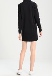 Robe longue noire à manches longues avec logo FILA au dos et texte accent blanc le long des manches ; design décontracté, longueur au genou, en tissu léger.