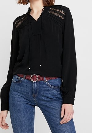 Zwarte blouse met lange mouwen en kanten details op de schouders, gecombineerd met blauwe spijkerbroek en een rode riem met een ronde zilveren gesp.