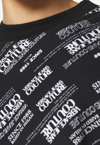 Černá bavlněná mikina se opakovaným potiskem "Versace Jeans Couture" a "Since 1988" v bílé barvě, přičemž text má různou orientaci.