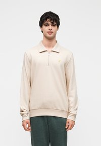 Junger Mann mit dunklem lockigem Haar, der einen beigefarbenen Pullover mit Reißverschluss am Kragen und dunkelgrüne Hose trägt, vor einem schlichten weißen Hintergrund steht.