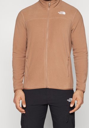 Mand iført en beige fleecejakke med lynlås og sorte bukser, begge med The North Face-logoer, stående mod en ensfarvet baggrund.