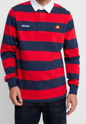 Mężczyzna ubrany w koszulkę rugby z długim rękawem w poziome paski czerwone i granatowe, z białym kołnierzem i naszywkami z logo ellesse na piersi.