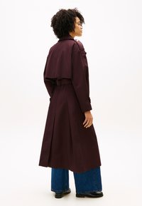 Μακρύ μπορντό παλτό τρανς με ζώνη στη μέση, πλαϊνές τσέπες και στυλάτο κάπα στο πίσω μέρος, συνδυασμένο με τζιν παντελόνια με φαρδιά μπατζάκια.