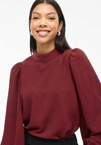 Blusa bordeaux a maniche lunghe con collo alto e maniche a sbuffo arricciate, realizzata in un tessuto testurizzato. Indossata con una vita nera.