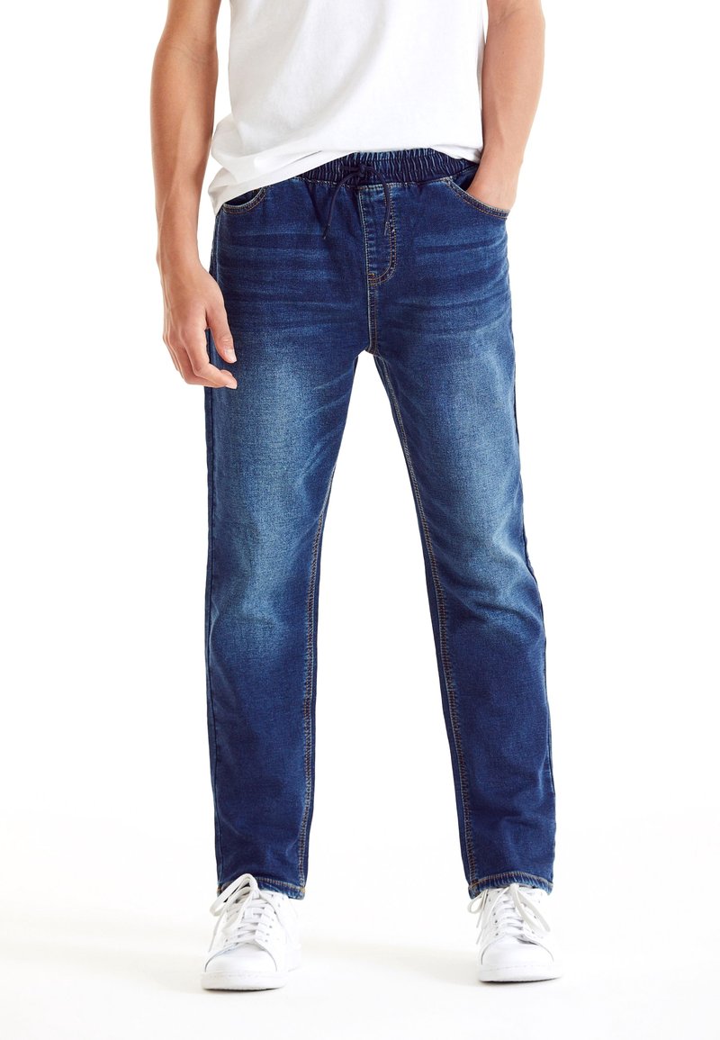 Homme portant un jean en denim à taille élastique bleu, des baskets blanches et une chemise blanche, debout avec une main dans la poche.