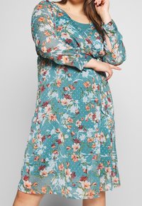 Robe florale en teal avec des manches longues et un col rond. Elle présente un tissu texturé avec de grandes fleurs multicolores et une coupe décontractée.