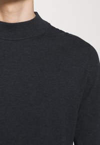 Dunkelgrauer Strickpullover mit hohem, geripptem Kragen. Der Stoff wirkt glatt und leicht strukturiert und hat eine taillierte Form ohne sichtbare Nähte.