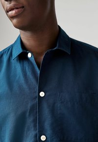 Camicia button-up blu scuro realizzata in tessuto strutturato, dotata di tasca, bottoni bianchi e un design del colletto classico.