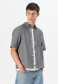 Short-sleeve, black and white checkered shirt with a button-up front, pocket detail, and a slight texture. Worn over a white T-shirt.