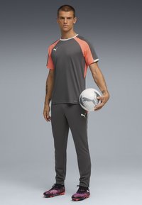 Camiseta deportiva gris con acentos en coral, combinada con pantalones grises ajustados. El modelo sostiene un balón de fútbol blanco con detalles en negro.