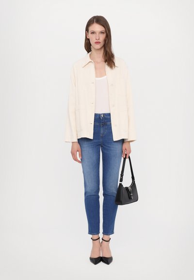 Lys beige overskjorte, hvit tanktop, blå smale jeans, svarte spisse hæler, holder en liten svart håndveske. Enkel og ren design.
