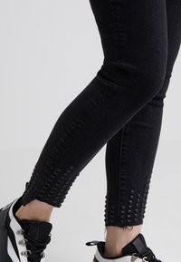 Black denim skinny jeans featuring a distressed hem and studded accents along the calves, paired with black and white sneakers.