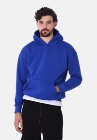 Felpa con cappuccio blu e tasca frontale, realizzata in tessuto morbido. Presenta una vestibilità comoda e polsini a coste. Indossata sopra una maglietta bianca.