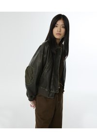 Chaqueta bomber de cuero verde oliva con parches acolchados en los codos, puños de canalé y un corte holgado. Combinada con pantalones de pana marrón.