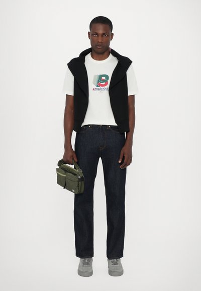 Svart hettevest over en hvit grafisk t-skjorte, mørke jeans, grå sneakers, holder en grønn bag. Enkelt design med kontrasterende farger.