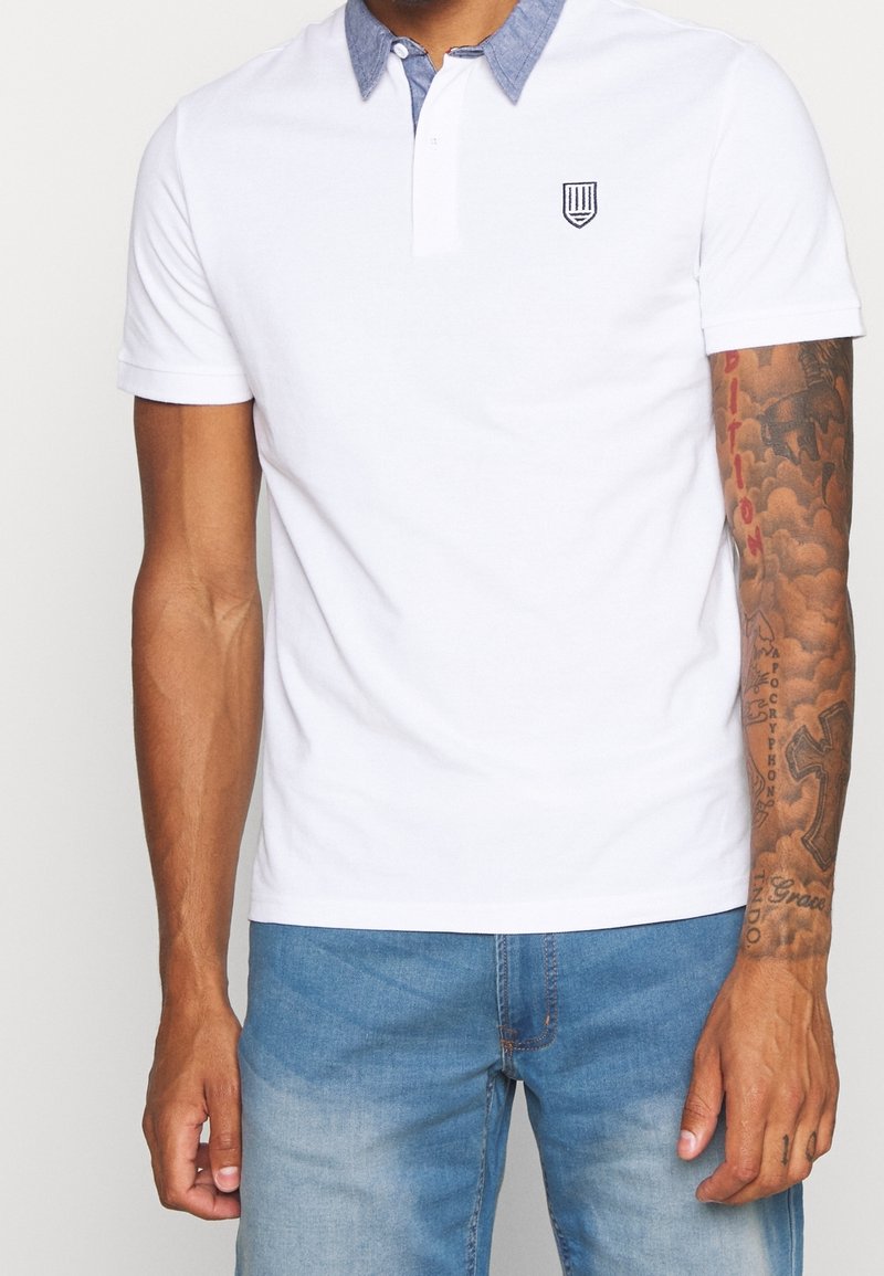 Homme portant un polo blanc à manches courtes avec col bleu et petit logo, jean bleu, tatouages couvrant l'avant-bras et la main droite.