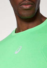 Lys grønn trenings T-skjorte med rund hals, laget av teksturert stoff, med en logo i lys gråfarge foran.