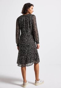 Femme portant une robe midi à manches longues noire et grise à motifs avec des poignets élastiques, associée à des baskets blanches, se tenant devant un fond blanc uni.
