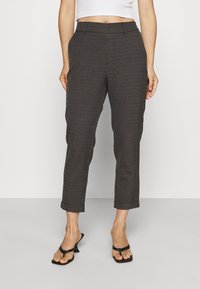 Kvinna i uppvikta rutiga mörka byxor, vit cropped top och svarta klacksandaler som står mot en enfärgad bakgrund.