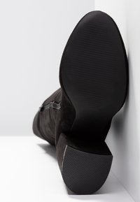 Botte en daim noir reposant sur le côté montrant une semelle lisse et un talon texturé sur fond blanc.