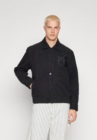 Sort uldjakke med knapper fortil, spidse kraver og sidelommer. Har et tekstureret broderet logo på brystet.
