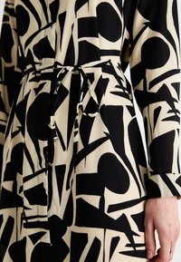 Abito nero e crema con motivi geometrici astratti, cintura annodata in vita, maniche lunghe e una texture di tessuto liscia.
