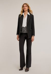 Schwarzer Blazer mit goldenen Reißverschlussdetails, weißes Hemd mit Knöpfen, schwarze Spitzenverzierung, maßgeschneiderte Hosen und schwarze Peeptoe-Absätze mit Netzakzenten.
