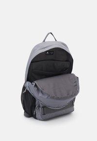 Grey backpack with a rounded top, front flap, and side mesh pocket. Features adjustable straps and a dark interior compartment.