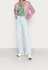 Veste bomber rose clair, haut fleuri vert, jeans taille haute bleu clair et baskets blanches avec des accents jaunes. Tenue décontractée.