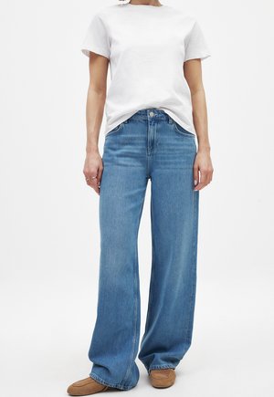 Jeans de mezclilla azul de pierna ancha con un lavado claro y un ligero desgaste, combinados con una camiseta blanca de algodón lisa y zapatillas planas marrones.