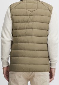 Olivgrüne Steppweste mit horizontalen Nähten, hohem Kragen, Eingrifftaschen und glatter Textur, getragen über einem hellen Pullover.